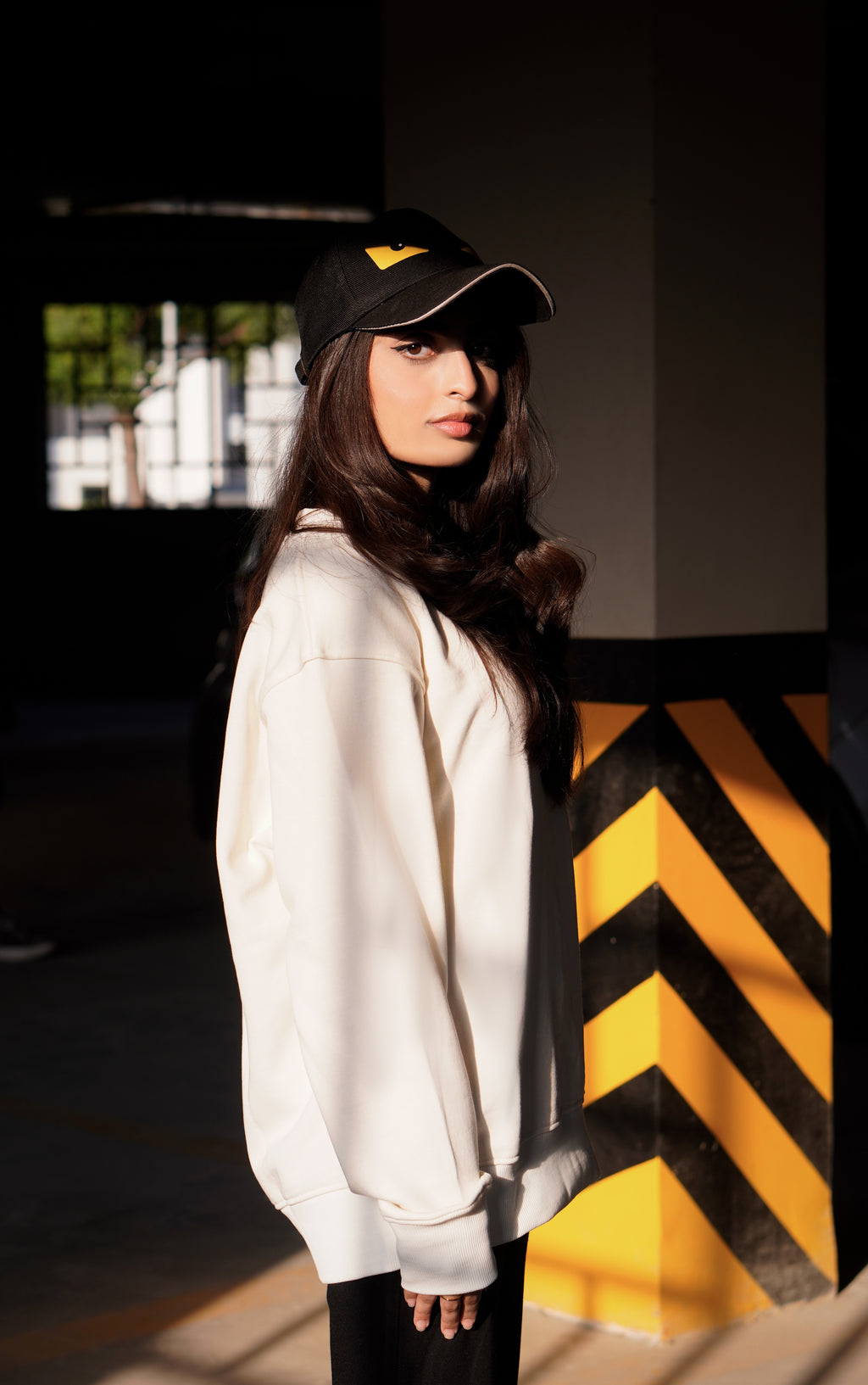 Person wearing a hoodie and black cap with yellow accents, standing in front of a black and yellow striped barrier.