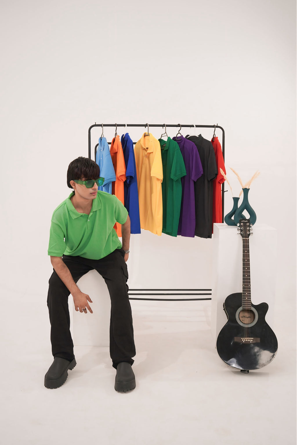 Person in a green POLO sitting next to a rack of colorful shirts and a guitar on a white background